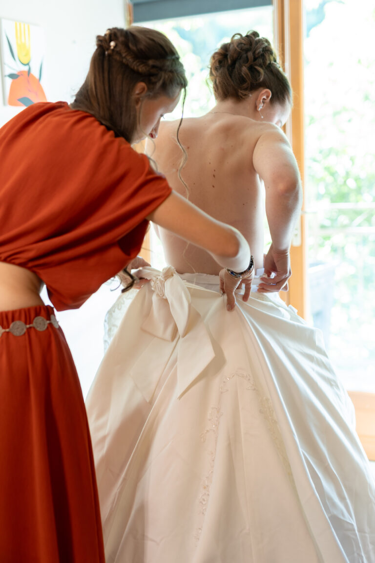 Une proche aide Jeanne à ajuster sa robe de mariée